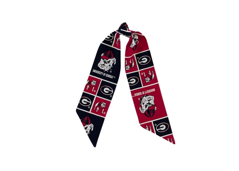 Puede incluir: Una bufanda larga y estrecha con un patr&oacute;n de cuadr&iacute;cula repetitivo en rojo, negro y blanco. El dise&ntilde;o incluye el texto University of Georgia, la mascota bulldog con gorra roja y el logotipo de la letra G impreso en la tela.