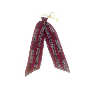 May include: A maroon ribbon keychain with the words "SOUTH CAROLINA" printed in white. The keychain has a gold-colored clasp and a small loop. The ribbon is folded in half, creating a loop.