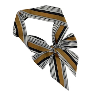 May include: A striped scarf in shades of gold, black, and white, tied in a bow. The scarf features a repeating pattern of vertical stripes. The accessory is made of a silky material.