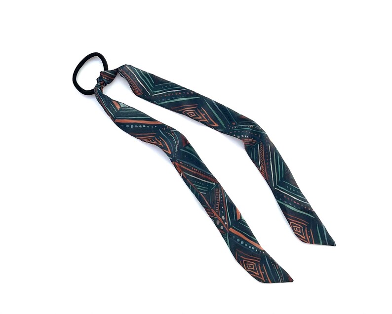 May include: A black hair tie with a long, patterned fabric tail. The fabric is dark green with a geometric pattern in shades of orange and brown.