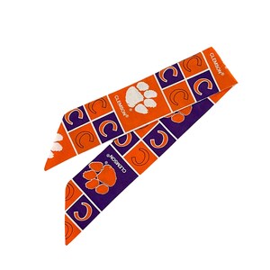 May include: Two fabric headbands in orange and purple, featuring a repeating pattern of the Clemson paw print and the letter "C" inside a square. The word "CLEMSON" is printed on the headband.