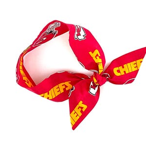 May include: A red fabric headband featuring a repeating pattern of the yellow text CHIEFS and the Kansas City Chiefs football team logo. The headband is tied into a knot on one side, creating a decorative bow effect with the printed fabric.