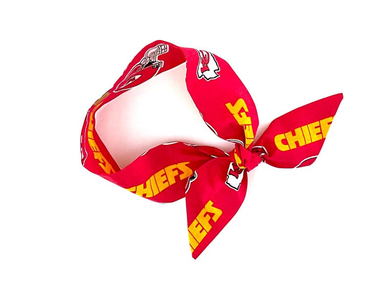 May include: A red fabric headband featuring a repeating pattern of the yellow text CHIEFS and the Kansas City Chiefs football team logo. The headband is tied into a knot on one side, creating a decorative bow effect with the printed fabric.