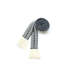 May include: A black and white woven belt with fringe tassels. The belt is rolled up and the fringe is visible.