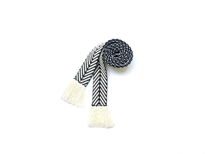 May include: A black and white woven belt with fringe tassels. The belt is rolled up and the fringe is visible.