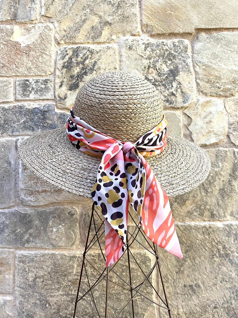 May include: A wide-brimmed straw hat with a pink, black, and gold patterned scarf tied around the crown.