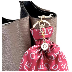 Pode incluir: Uma bolsa de couro taupe com uma al&ccedil;a preta e um chaveiro dourado. Um len&ccedil;o vermelho com texto branco est&aacute; preso ao chaveiro, junto com um pequeno pingente redondo com a letra "O". A bolsa tem uma superf&iacute;cie texturizada.