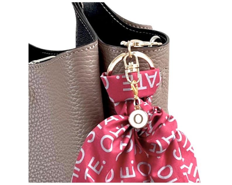 Pode incluir: Uma bolsa de couro taupe com uma al&ccedil;a preta e um chaveiro dourado. Um len&ccedil;o vermelho com texto branco est&aacute; preso ao chaveiro, junto com um pequeno pingente redondo com a letra "O". A bolsa tem uma superf&iacute;cie texturizada.