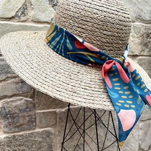 May include: A wide-brimmed straw hat with a colourful patterned fabric band tied around the crown. The band features a teal, pink and yellow abstract design.