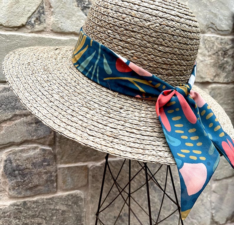May include: A wide-brimmed straw hat with a colourful patterned fabric band tied around the crown. The band features a teal, pink and yellow abstract design.