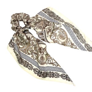 May include: A cream-colored scrunchie with attached fabric tails. The fabric features a baroque-style pattern in shades of brown and black, with a Greek key border. The scrunchie is designed for hair styling.