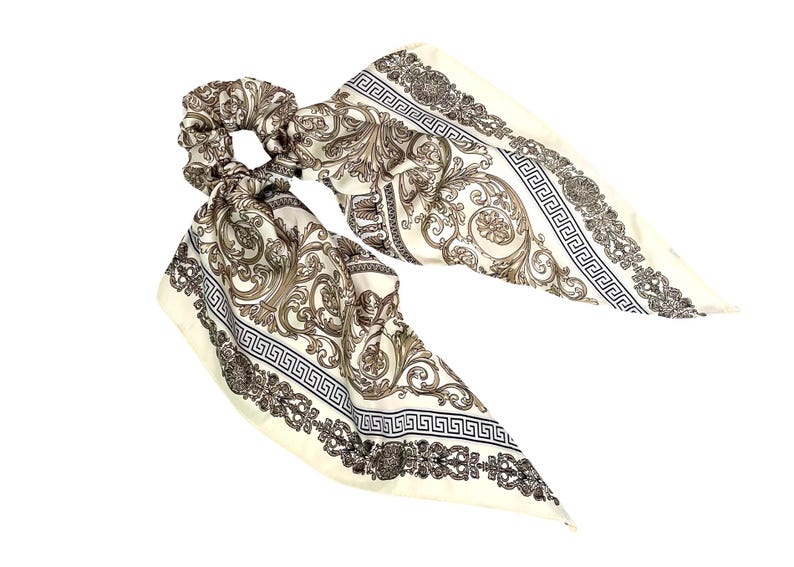 May include: A cream-colored scrunchie with attached fabric tails. The fabric features a baroque-style pattern in shades of brown and black, with a Greek key border. The scrunchie is designed for hair styling.