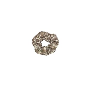 May include: A beige and brown patterned scrunchie. The fabric features a repeating ornate design in shades of beige and brown. The scrunchie is circular and appears to be made of a soft, silky material. This hair accessory is suitable for various hairstyles.