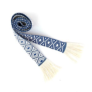 May include: A white and blue woven belt with fringe tassels. The belt has a geometric diamond pattern.