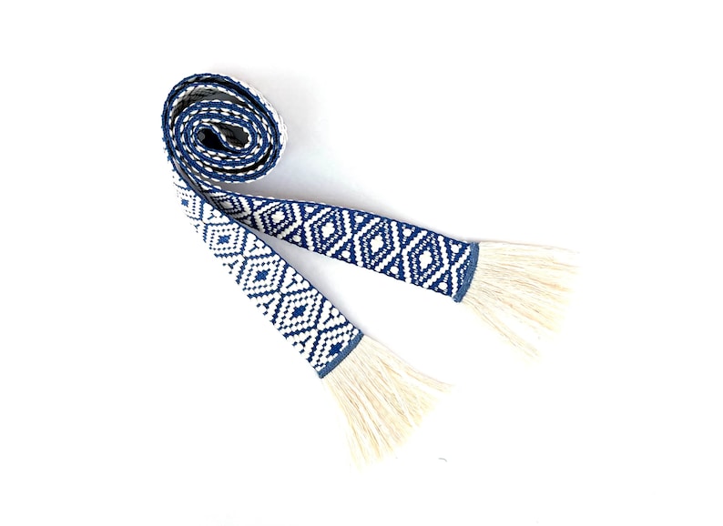 May include: A white and blue woven belt with fringe tassels. The belt has a geometric diamond pattern.