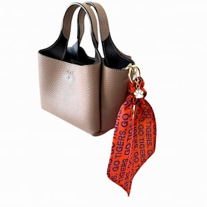 May include: A taupe-colored handbag with two handles and a black interior. A decorative orange scarf with the text "GO TIGERS" is attached to the bag with a gold-colored charm. The scarf has a paw print charm.