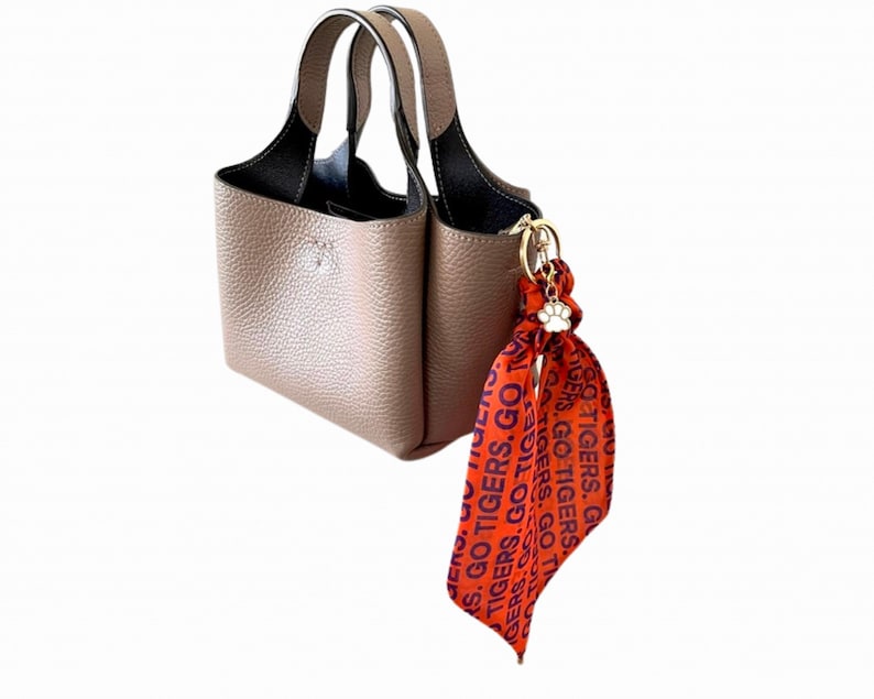 May include: A taupe-colored handbag with two handles and a black interior. A decorative orange scarf with the text "GO TIGERS" is attached to the bag with a gold-colored charm. The scarf has a paw print charm.