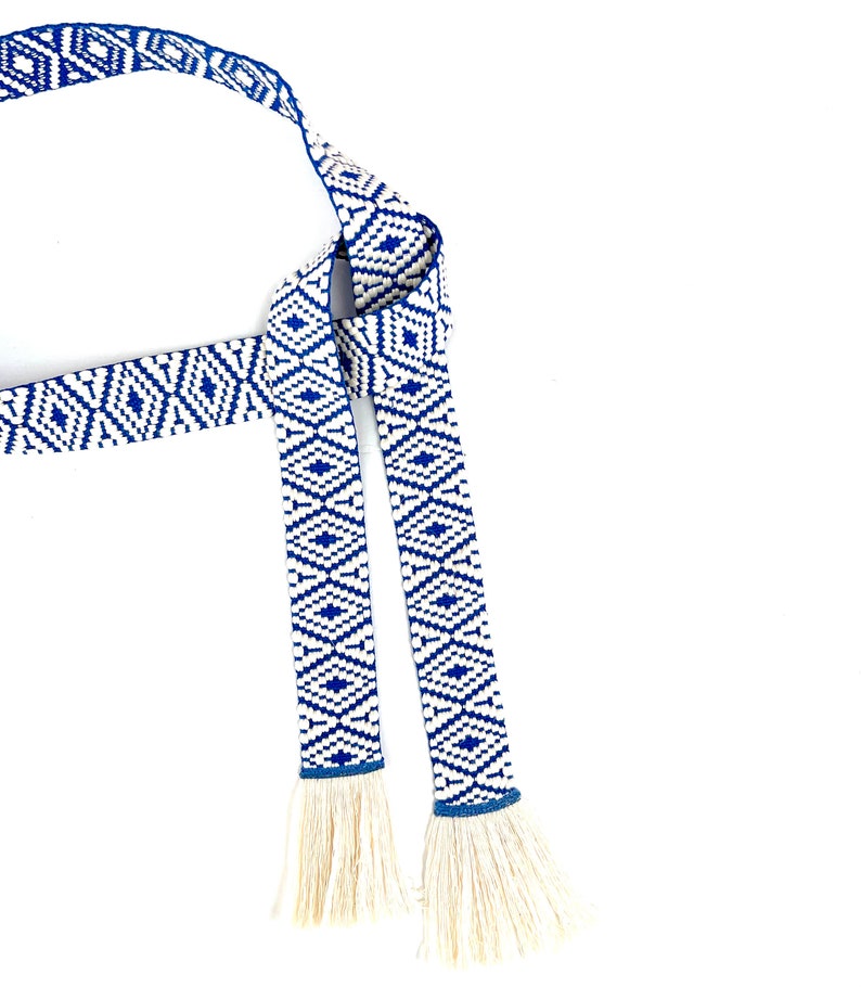 May include: A blue and white woven belt with tassels. The belt has a geometric pattern and is tied in a knot.