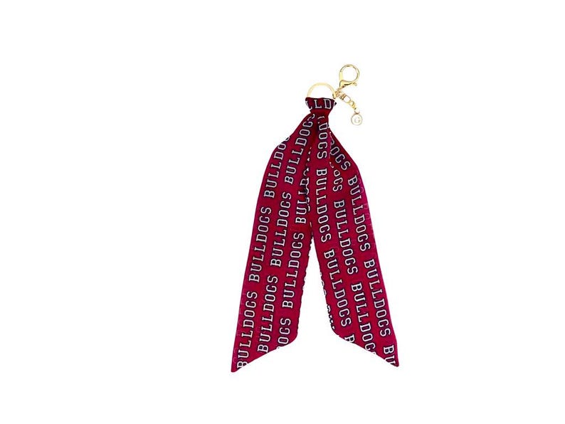 May include: A maroon and white Bulldogs keychain with a gold-colored clasp. The keychain features a long, ribbon-like design with the word "BULLDOGS" printed repeatedly in white.