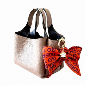 May include: A taupe-colored handbag with a black interior and matching handles. A decorative orange bow with the text "GO TIGERS" is attached with a gold-colored charm and a white paw print.
