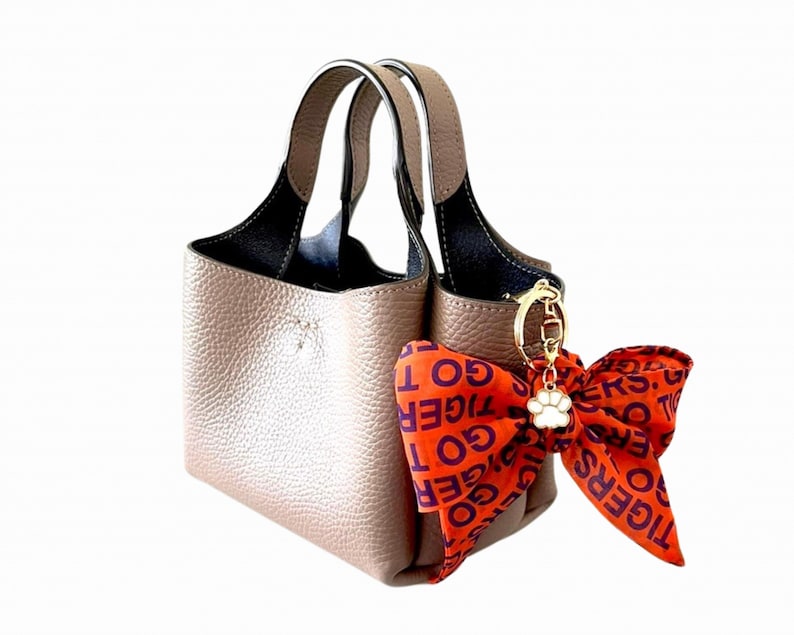 May include: A taupe-colored handbag with a black interior and matching handles. A decorative orange bow with the text "GO TIGERS" is attached with a gold-colored charm and a white paw print.