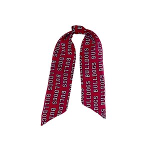 Puede incluir: Una cinta o pa&ntilde;uelo largo y estrecho de tela roja con un patr&oacute;n de texto blanco repetitivo que dice BULLDOGS. El accesorio est&aacute; atado con un nudo simple en la parte superior, creando dos extremos largos y afilados que cuelgan.