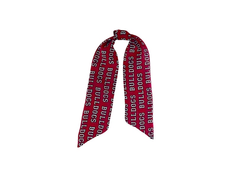 Puede incluir: Una cinta o pa&ntilde;uelo largo y estrecho de tela roja con un patr&oacute;n de texto blanco repetitivo que dice BULLDOGS. El accesorio est&aacute; atado con un nudo simple en la parte superior, creando dos extremos largos y afilados que cuelgan.