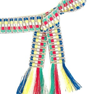 Puede incluir: Un cintur&oacute;n de tela tejida con un patr&oacute;n geom&eacute;trico repetitivo en rojo, azul, amarillo, verde y blanco. El cintur&oacute;n est&aacute; anudado y rematado con largos flecos multicolores en los extremos, que incorporan la misma paleta de colores vivos.