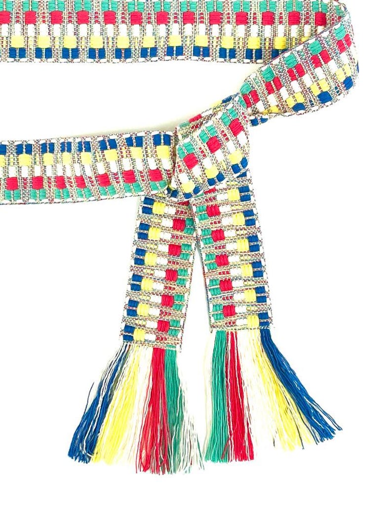 Puede incluir: Un cintur&oacute;n de tela tejida con un patr&oacute;n geom&eacute;trico repetitivo en rojo, azul, amarillo, verde y blanco. El cintur&oacute;n est&aacute; anudado y rematado con largos flecos multicolores en los extremos, que incorporan la misma paleta de colores vivos.
