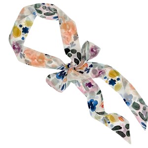 May include: A floral patterned scarf with a bow. The scarf features a watercolor design with flowers in shades of pink, yellow, blue, and green on a white background. The scarf is tied in a bow.