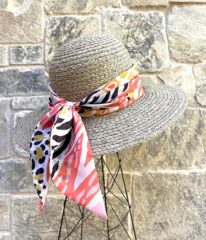 May include: A wide-brimmed straw hat with a colorful patterned scarf tied around the crown. The scarf features pink, black, white, and yellow abstract shapes.