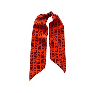 May include: Orange fabric hair scarf with the text "GO TIGERS" in black repeated vertically. The scarf is tied at the top, with two long, pointed ends hanging down.