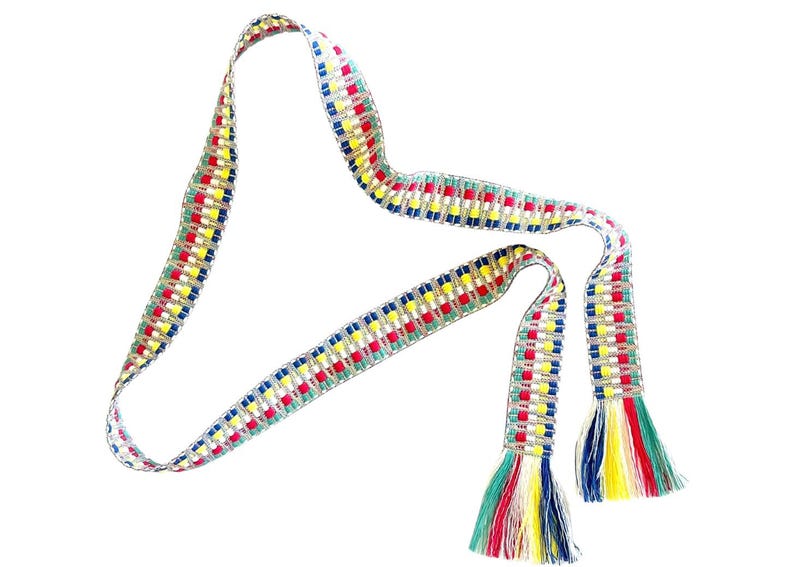 Puede incluir: Un cintur&oacute;n o faja tejida larga y estrecha con un patr&oacute;n geom&eacute;trico repetitivo en hilos rojos, amarillos, azules y blancos. Los extremos del cintur&oacute;n est&aacute;n terminados con borlas de flecos multicolores en tonos verde, rojo, amarillo, blanco y azul.