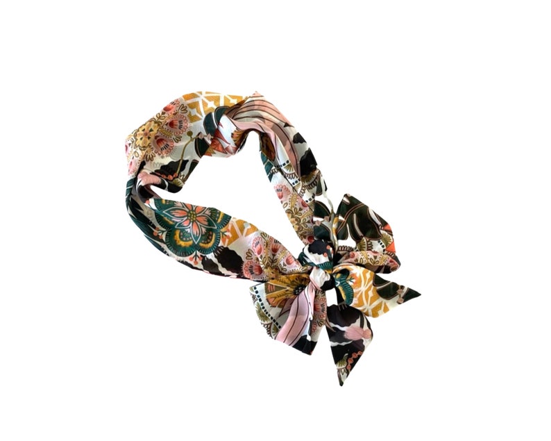 May include: A fabric headband featuring a vibrant, multi-colored floral and geometric pattern in shades of mustard yellow, deep green, black, and soft pink. The accessory is tied into a decorative bow knot on one side, creating a structured loop.