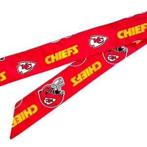 May include: A long, red fabric hair ribbon featuring a repeating pattern of the yellow text CHIEFS, white and red football helmet graphics, and white arrowhead logos with the letters KC printed in red on the vibrant red background.