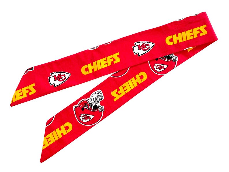 May include: A long, red fabric hair ribbon featuring a repeating pattern of the yellow text CHIEFS, white and red football helmet graphics, and white arrowhead logos with the letters KC printed in red on the vibrant red background.