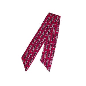 Puede incluir: Una cinta o pa&ntilde;uelo largo y estrecho de color magenta que presenta el texto blanco repetido "BULLDOGS" impreso en una fuente negrita sin remates. El accesorio tiene extremos puntiagudos y angulados con un patr&oacute;n constante en toda su longitud.