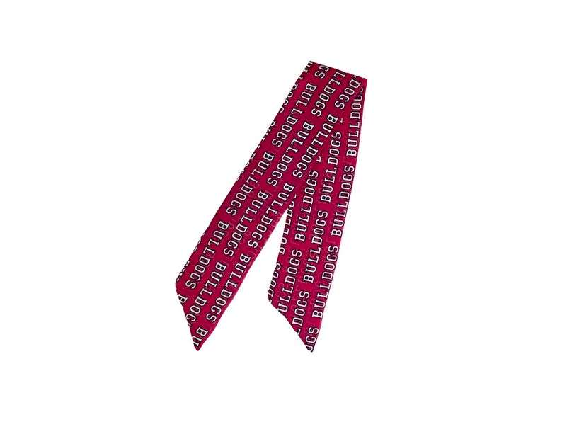 Puede incluir: Una cinta o pa&ntilde;uelo largo y estrecho de color magenta que presenta el texto blanco repetido "BULLDOGS" impreso en una fuente negrita sin remates. El accesorio tiene extremos puntiagudos y angulados con un patr&oacute;n constante en toda su longitud.