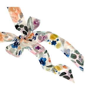 May include: A floral patterned scarf with a bow. The scarf features watercolor-style flowers in shades of orange, blue, purple, and green on a white background. The scarf is tied in a bow.