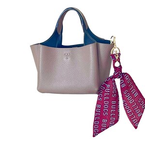 Puede incluir: Un bolso de mano color topo con superficie texturizada, que cuenta con dos asas azul oscuro y un interior azul oscuro. Un lazo granate con el texto blanco repetido "BULLDOGS" est&aacute; sujeto al lateral del bolso con un cierre de metal dorado.