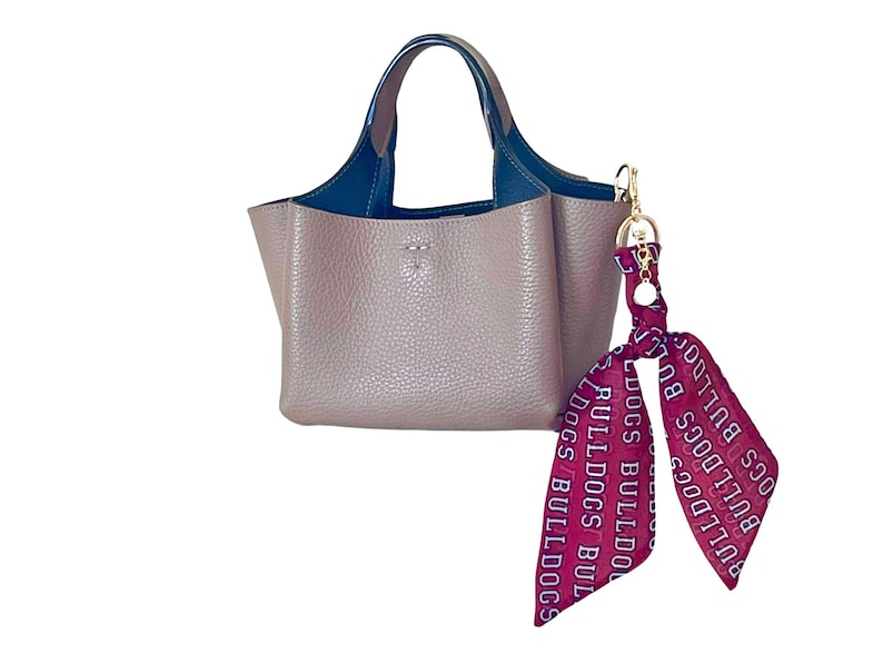 Puede incluir: Un bolso de mano color topo con superficie texturizada, que cuenta con dos asas azul oscuro y un interior azul oscuro. Un lazo granate con el texto blanco repetido "BULLDOGS" est&aacute; sujeto al lateral del bolso con un cierre de metal dorado.