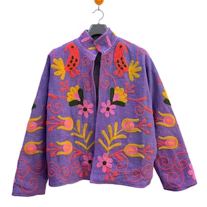 Peut inclure: Veste violette ornée de broderies florales colorées. La veste présente un col montant et est ornée d'oiseaux brodés, de fleurs et de volutes dans les tons de rouge, jaune, rose et vert. Accrochée à un cintre noir.
