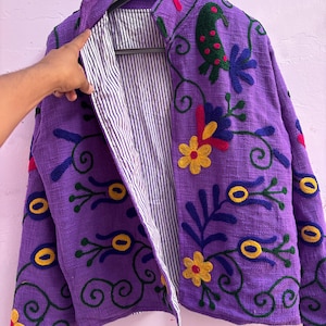 Peut inclure: Veste violette avec broderies florales jaunes, vertes, rouges et bleues. La veste a une doublure rayée et est suspendue à un cintre noir. La broderie présente des motifs de fleurs et de feuilles stylisés.