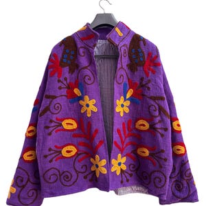 Peut inclure: Une veste violette avec des broderies florales colorées. La veste a un col montant et est doublée d'un tissu rayé. La broderie comprend des fleurs et des feuilles rouges, jaunes et marron, créant un design vibrant.