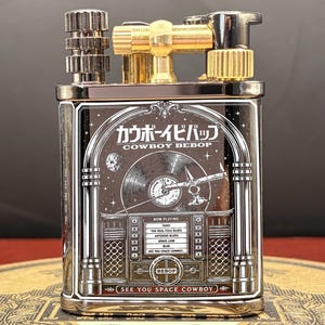 Op de afbeelding: Een zilveren en gouden metalen aansteker met de Japanse en Engelse tekst "COWBOY BEBOP". De aansteker heeft een gedetailleerde afbeelding van een jukebox met een plaat en de woorden "NOW PLAYING" en "SEE YOU SPACE COWBOY".