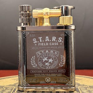 May include: A silver and gold metal lighter featuring the text "S.T.A.R.S. FIELD CASE" and "RACCOON CITY POLICE DEPT." The rectangular lighter has a hinged top and a textured wheel. It is a collectible item, with a serial number and unit issue details.