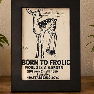 May include: A black framed print featuring a black and white illustration of a spotted fawn. The text reads: BORN TO FROLIC, WORLD IS A GARDEN, Love Em All 1989, I am alive, 410,737,684,530 JOYS. The print has a cream-coloured background.