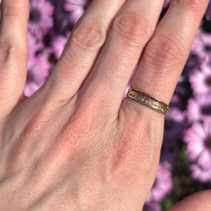 May include: A gold ring with a double band design. The top band features a row of small red gemstones, while the bottom band has a row of clear gemstones. The ring is worn on a finger against a blurred floral background.