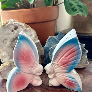 May include: A pair of ceramic salt and pepper shakers shaped like butterflies. Each shaker features a glossy white base with gradient wings colored in soft pink and bright blue. The top of one shaker has small holes for dispensing seasoning.