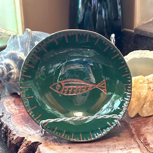 May include: A small, round, dark green ceramic dish featuring a central rust-colored fish illustration and a decorative rust-colored ruler-style border around the rim. The dish is held upright by a silver-toned twisted wire display stand.